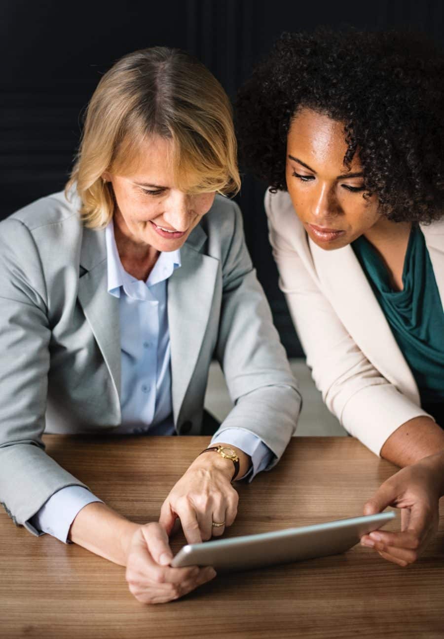Two women in professional attire discuss something on a tablet while seated at a wooden table.
