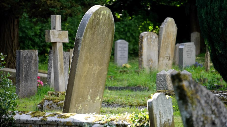 A cluster of old, weathered gravestones in a lush, green cemetery. Some headstones are upright while others are tilted or covered in moss.
