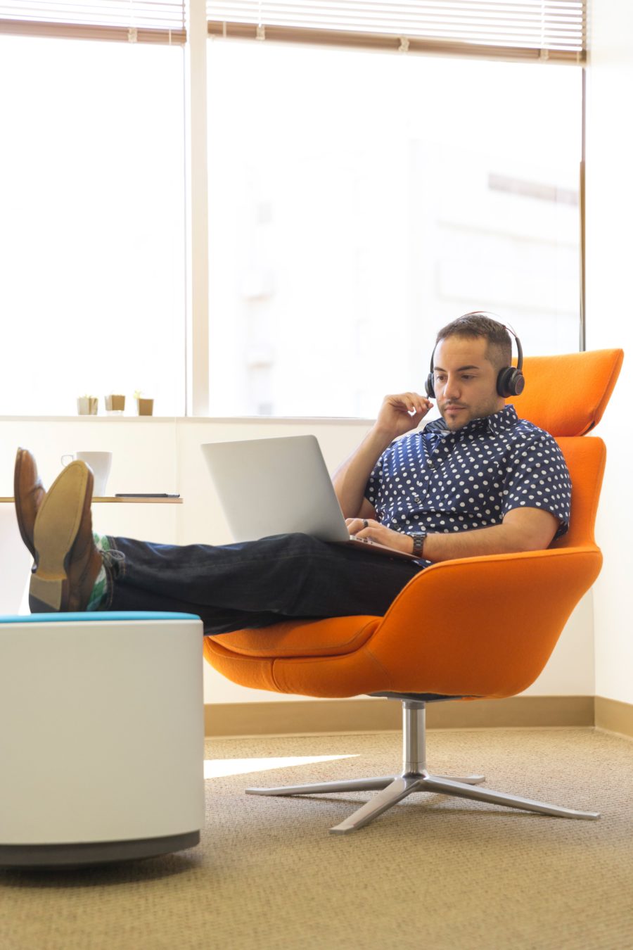Man sitting in an orange chair with feet up, wearing headphones, and working on a laptop in a bright room.