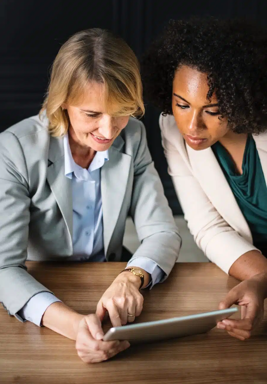 Two women in professional attire discuss something on a tablet while seated at a wooden table.