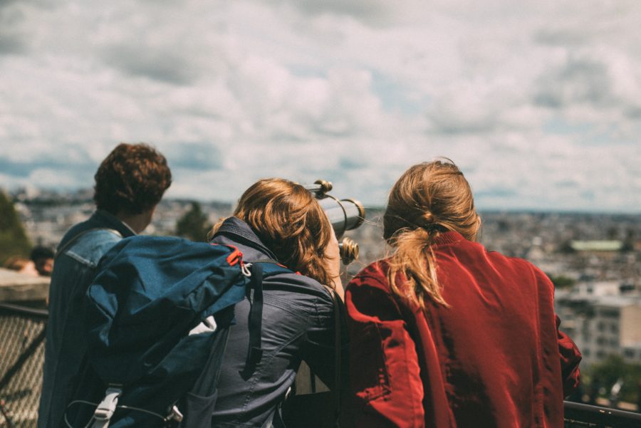 Three people, two with long blond hair facing away from the camera, use a public telescope to look at a cityscape on a cloudy day.