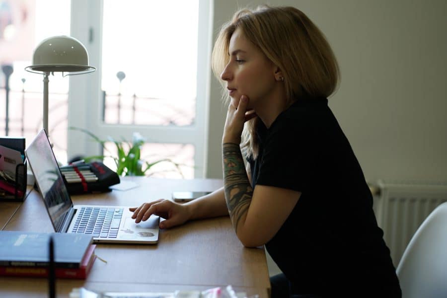 A person with long hair and a tattoo on their left arm works on a laptop at a desk in a well-lit room.