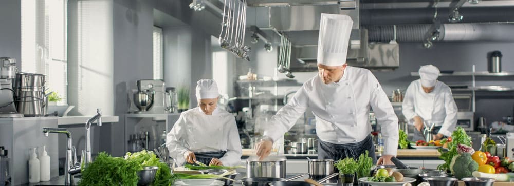 Three chefs in a professional kitchen prepare various dishes using fresh vegetables.