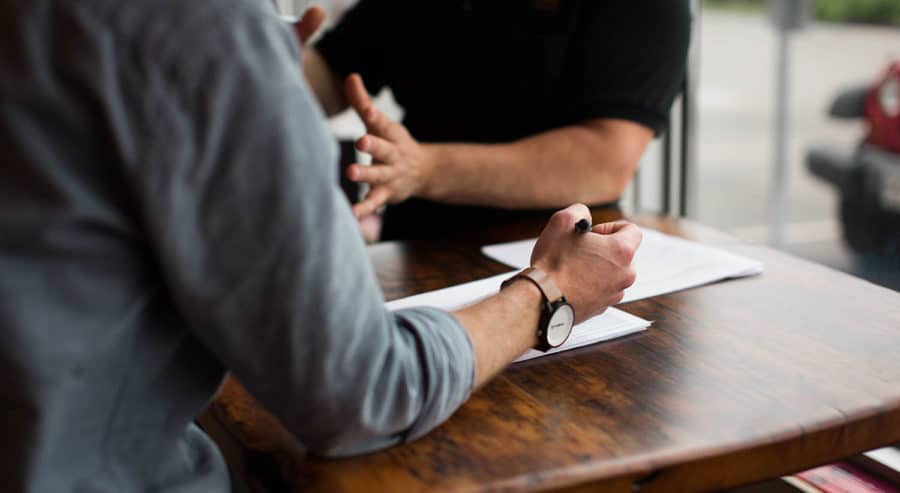 Two people sitting across from each other at a wooden table, discussing documents. One person is holding a pen, and papers are spread out on the table.