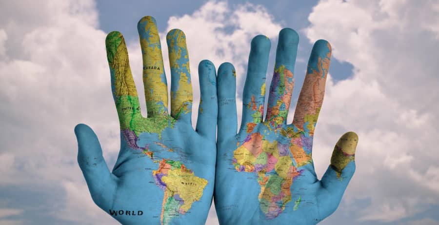 Two hands are painted with a world map, displaying different continents and countries. The background shows a cloudy sky.