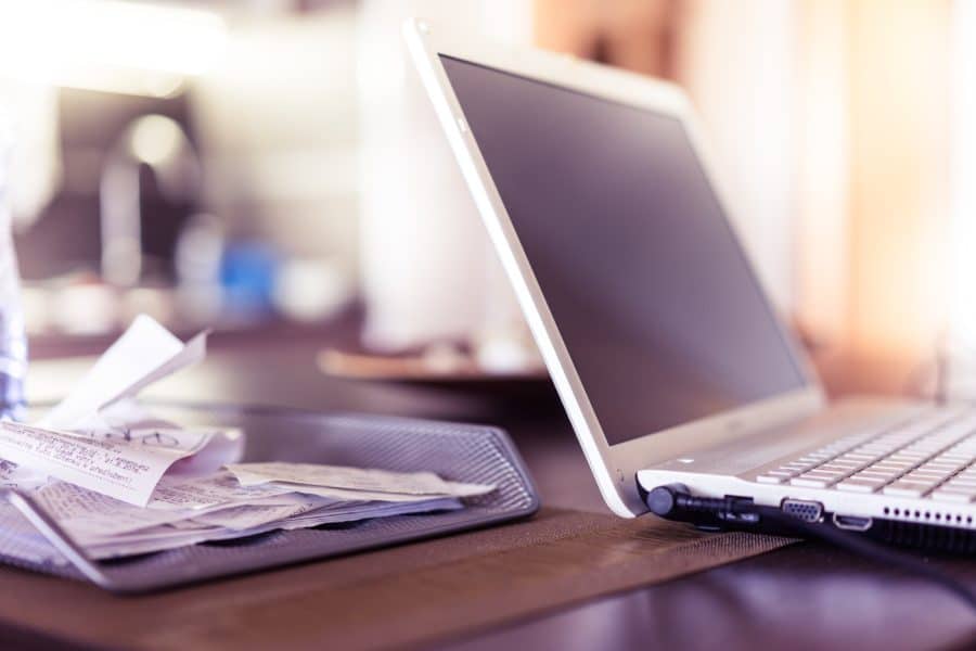 A laptop computer is open on a table next to a mesh tray filled with various receipts and papers, likely detailing staff expenses. The background is blurred.