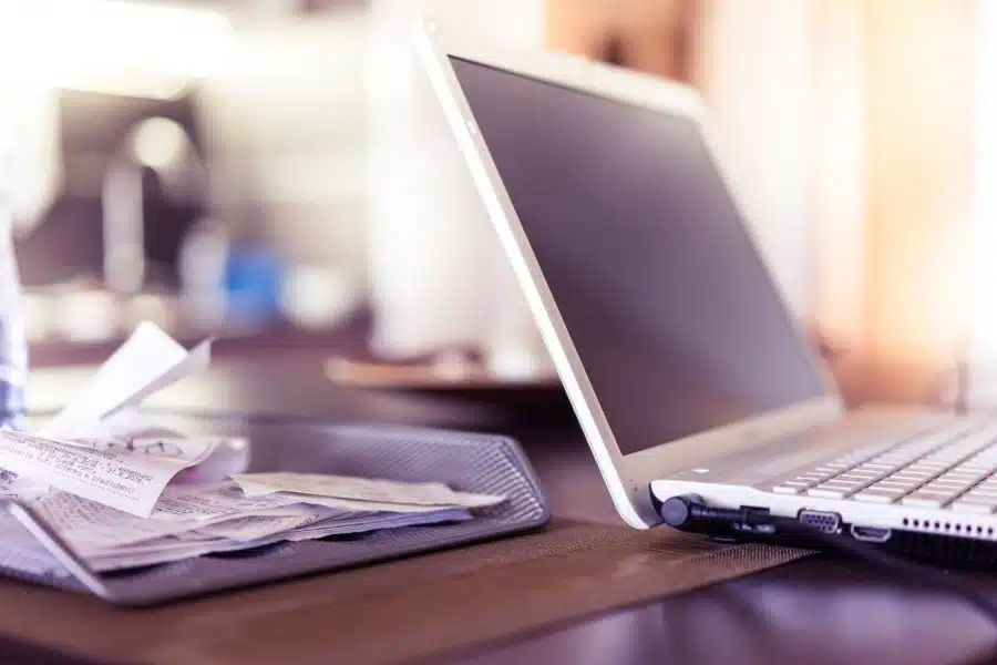 A laptop computer is open on a table next to a mesh tray filled with various receipts and papers, likely detailing staff expenses. The background is blurred.