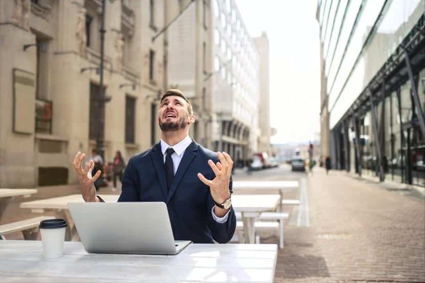 A man in a suit sits at an outdoor table with a laptop and coffee cup, looking up with a frustrated expression, hands raised, likely dealing with a problematic browser.