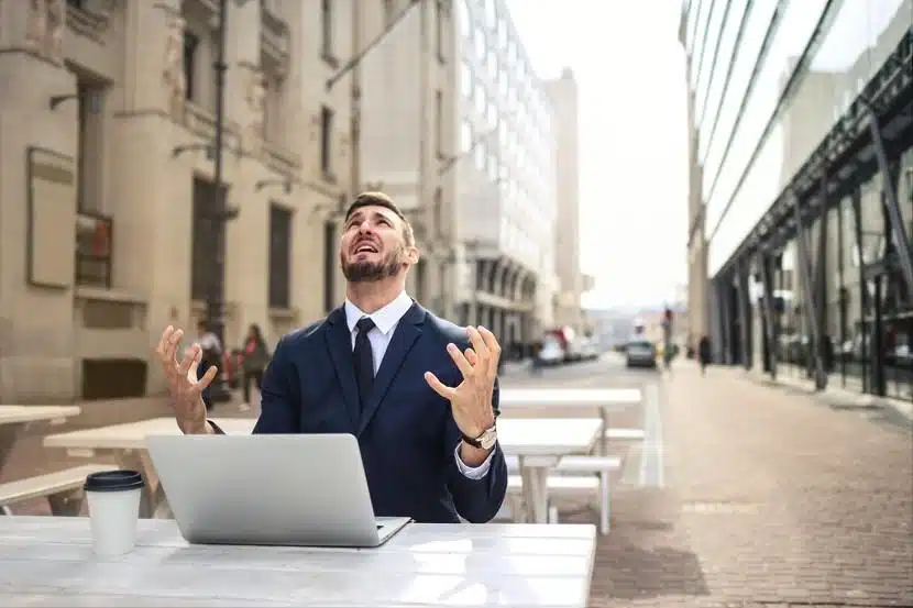 A man in a suit sits at an outdoor table with a laptop and coffee cup, looking up with a frustrated expression, hands raised, likely dealing with a problematic browser.