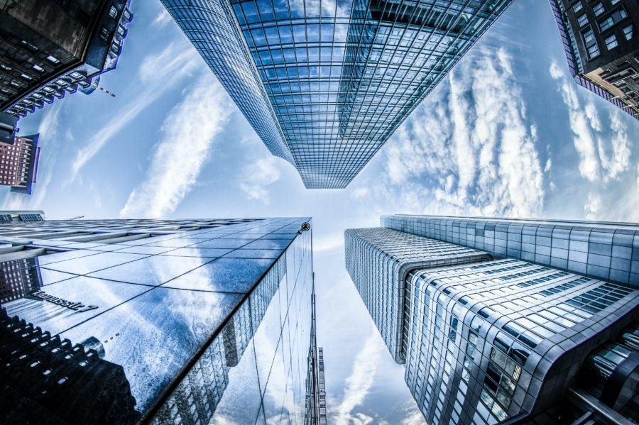 A fisheye lens view of modern skyscrapers with glass facades, reflecting the cloud-dotted blue sky, taken from a low angle looking up.