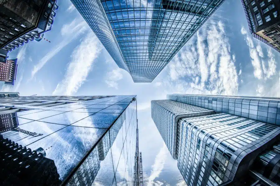A fisheye lens view of modern skyscrapers with glass facades, reflecting the cloud-dotted blue sky, taken from a low angle looking up.
