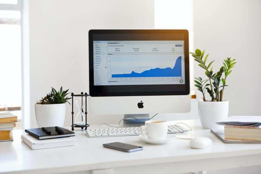 A computer on a white desk displays a graph on its screen, signaling it's time to get back to work. The desk also holds plants, notebooks, a smartphone, a coffee cup, and a mouse.
