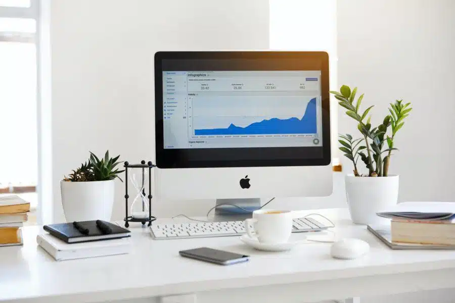 A computer on a white desk displays a graph on its screen, signaling it's time to get back to work. The desk also holds plants, notebooks, a smartphone, a coffee cup, and a mouse.
