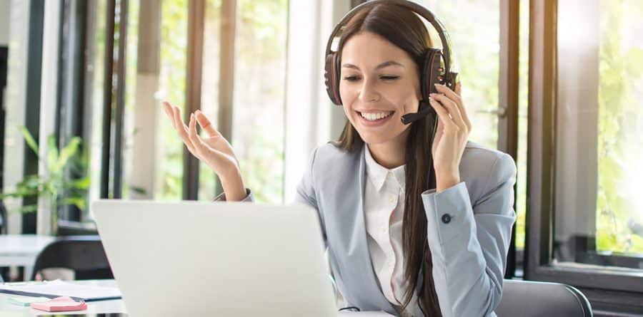 A woman wearing one of the best headsets and a gray blazer smiles while engaging in a video call on a laptop. She gestures with one hand. The background includes windows with greenery outside.