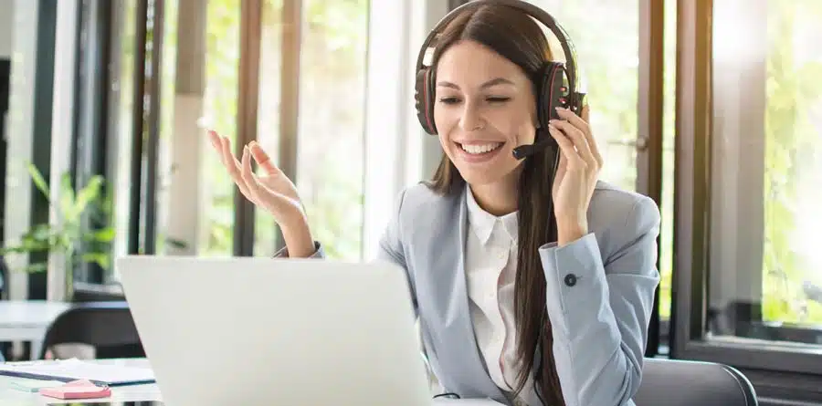 A woman wearing one of the best headsets and a gray blazer smiles while engaging in a video call on a laptop. She gestures with one hand. The background includes windows with greenery outside.