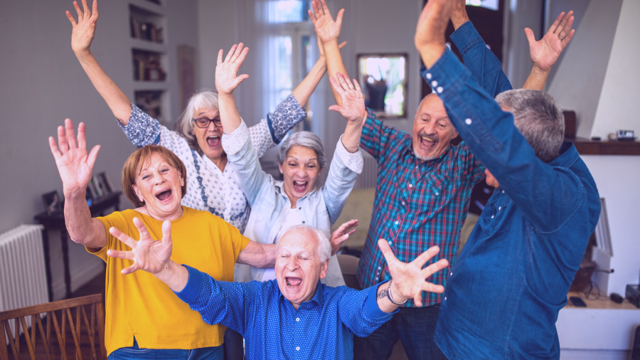 A group of elderly people from local care homes with their arms raised, smiling and cheering enthusiastically in a brightly lit indoor setting.
