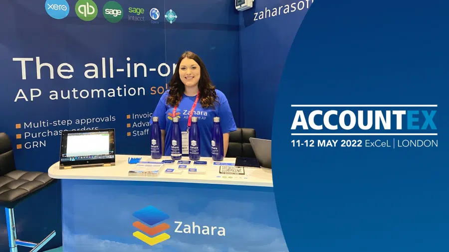 A woman stands at the Zahara booth at ACCOUNTS 2022, ExCeL London. The Accountex booth highlights AP automation solutions with various promotional materials on display, including a laptop, pamphlets, and water bottles.