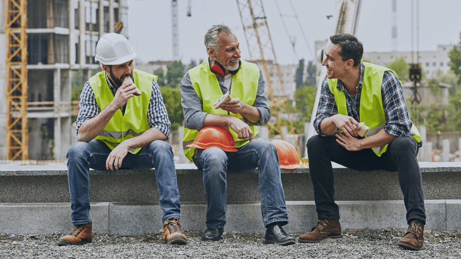 Three construction workers wearing safety vests and helmets take a break, sitting on concrete with a construction site in the background. One is eating, while two are engaged in conversation, smiling, possibly discussing the best way to handle processing invoices.